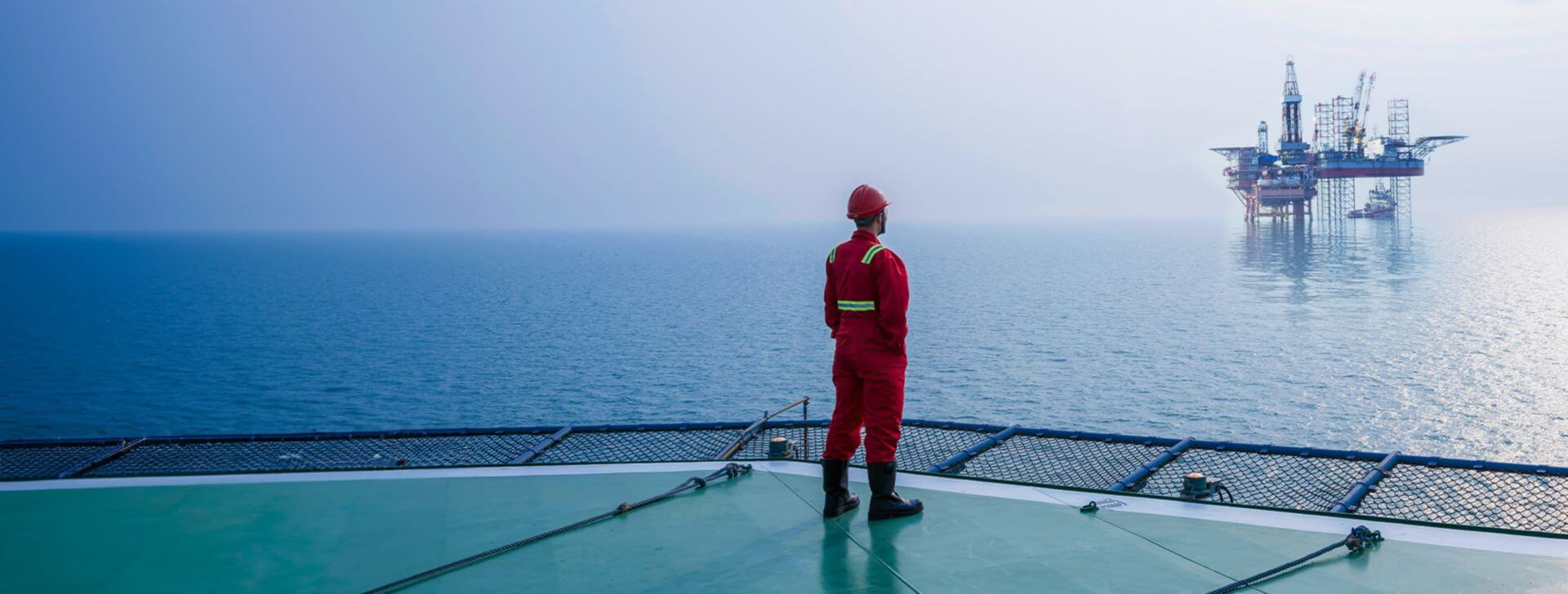 Offshore worker in safety gear standing on a vessel deck facing an oil drilling platform at sea.