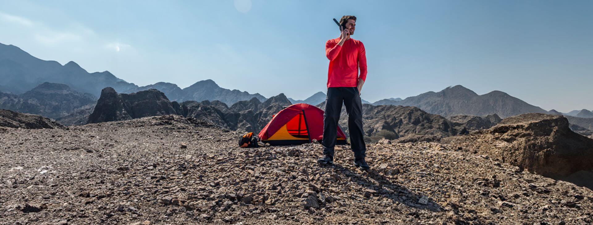 Man near a tent in a rocky mountain landscape, holding a satellite phone under a clear blue sky.