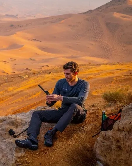 Man sitting on a rocky desert hillside using a satellite phone, with dunes and mountains in the background.
