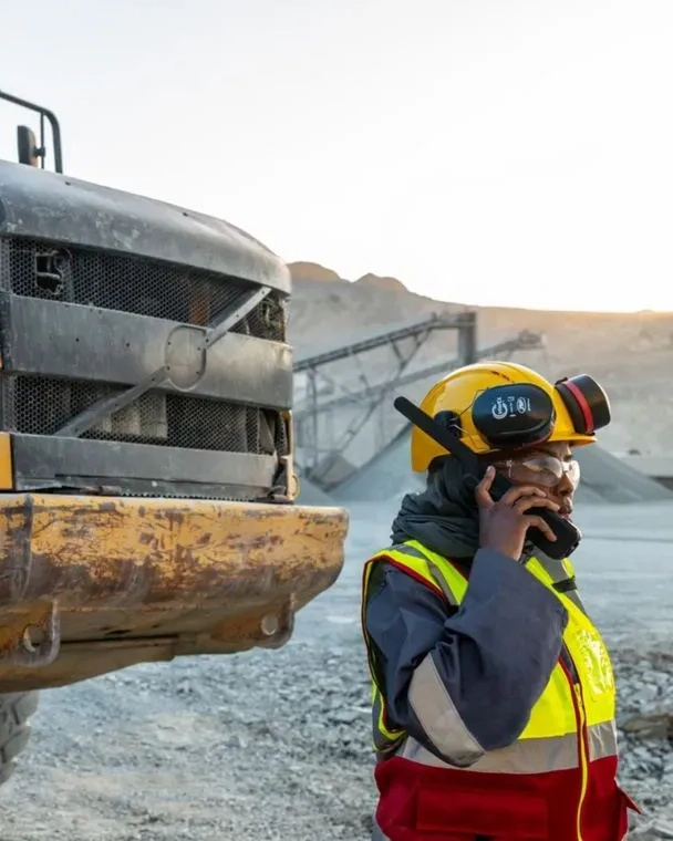Construction worker in safety gear using a satellite phone beside heavy machinery at a quarry site.