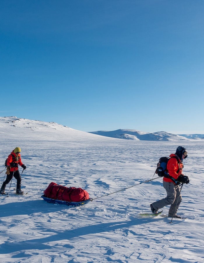 Two explorers pulling sleds across a snowy arctic landscape under clear skies.
