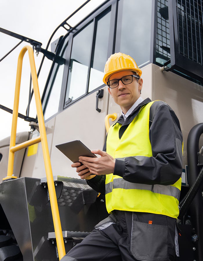 Construction worker in hard hat and safety vest holding a tablet near heavy machinery.