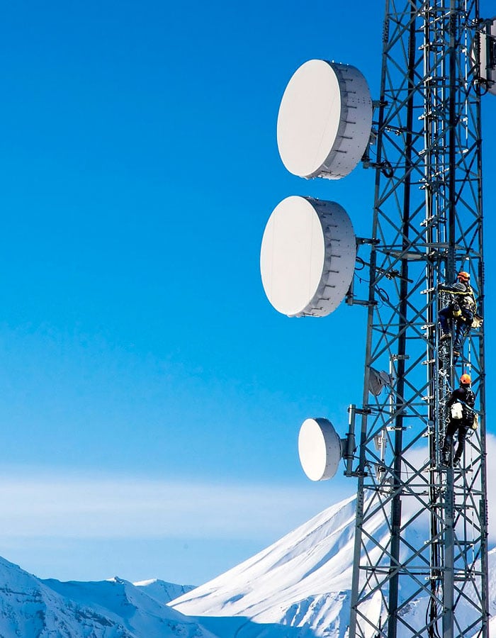 Workers climbing a tall communications tower in a snowy mountain landscape.