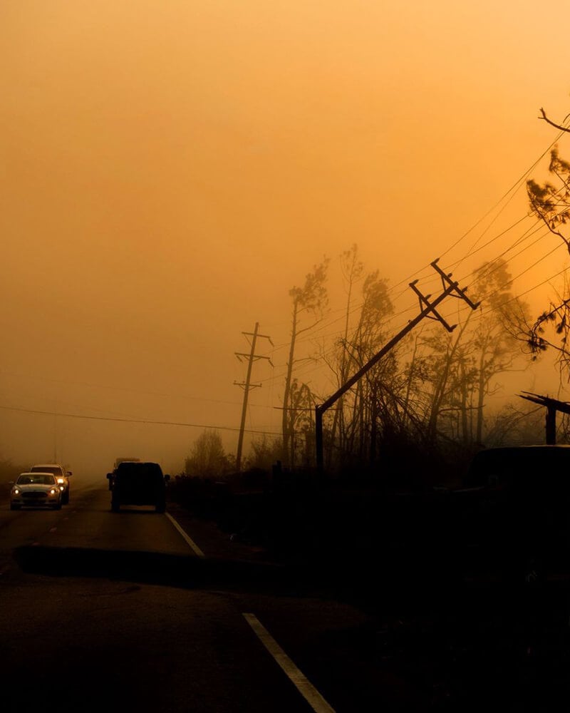 Smoke-filled roadway with cars driving under an orange sky, damaged power lines and trees silhouetted along the roadside.