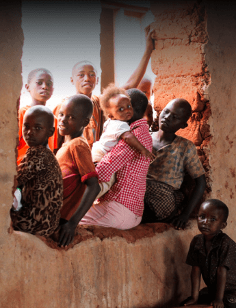 Group of children and a woman gathered in a rough brick window opening, looking toward the camera in a rural setting.